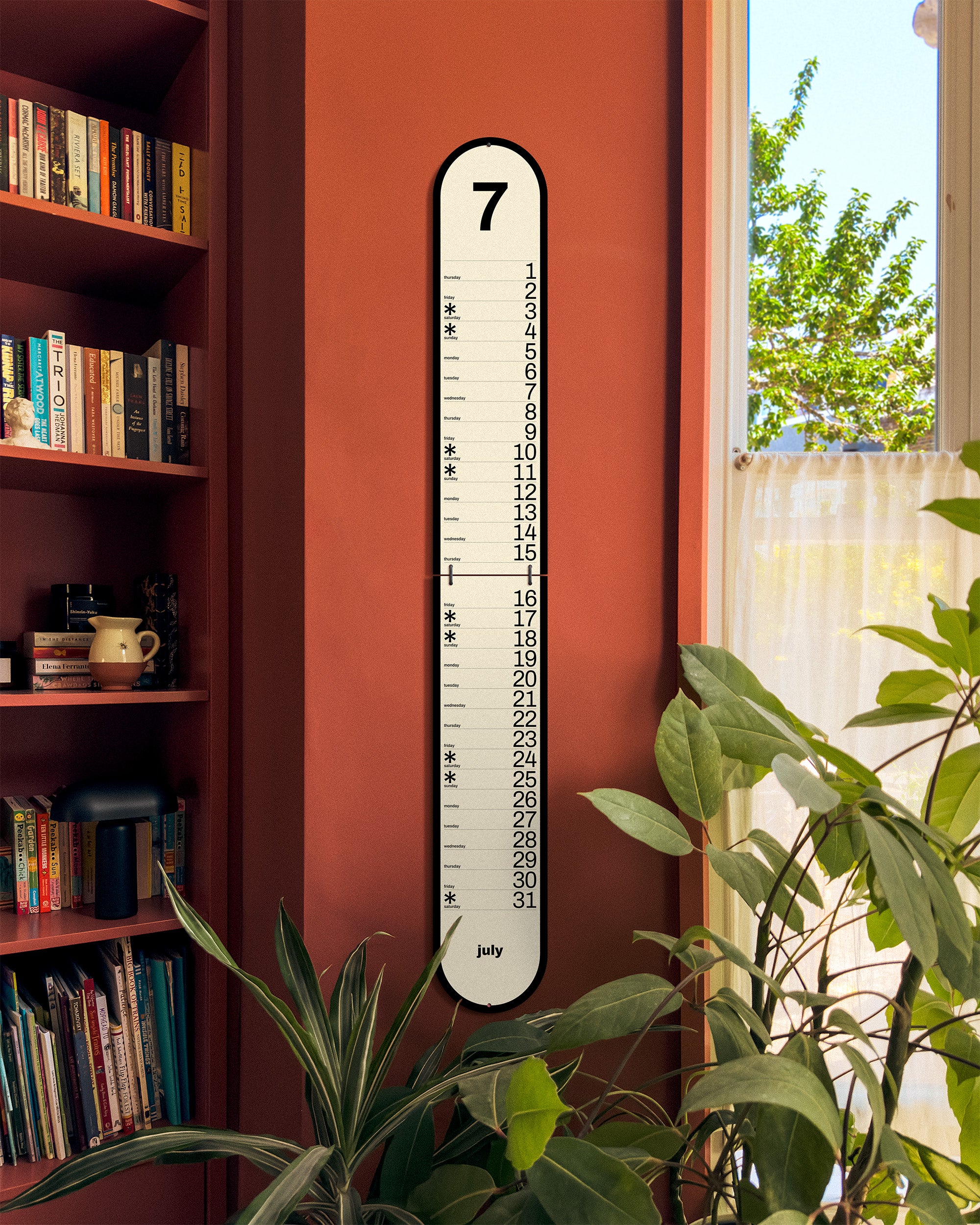 Wall-mounted Obok calendar against a red wall with plants and books.