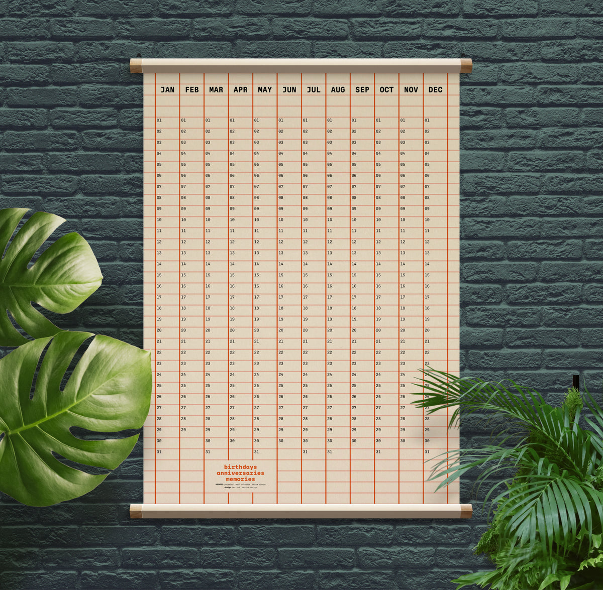 A vertical monthly calendar poster with a beige grid and wooden frame, mounted against a dark green brick wall with green plants in the foreground.