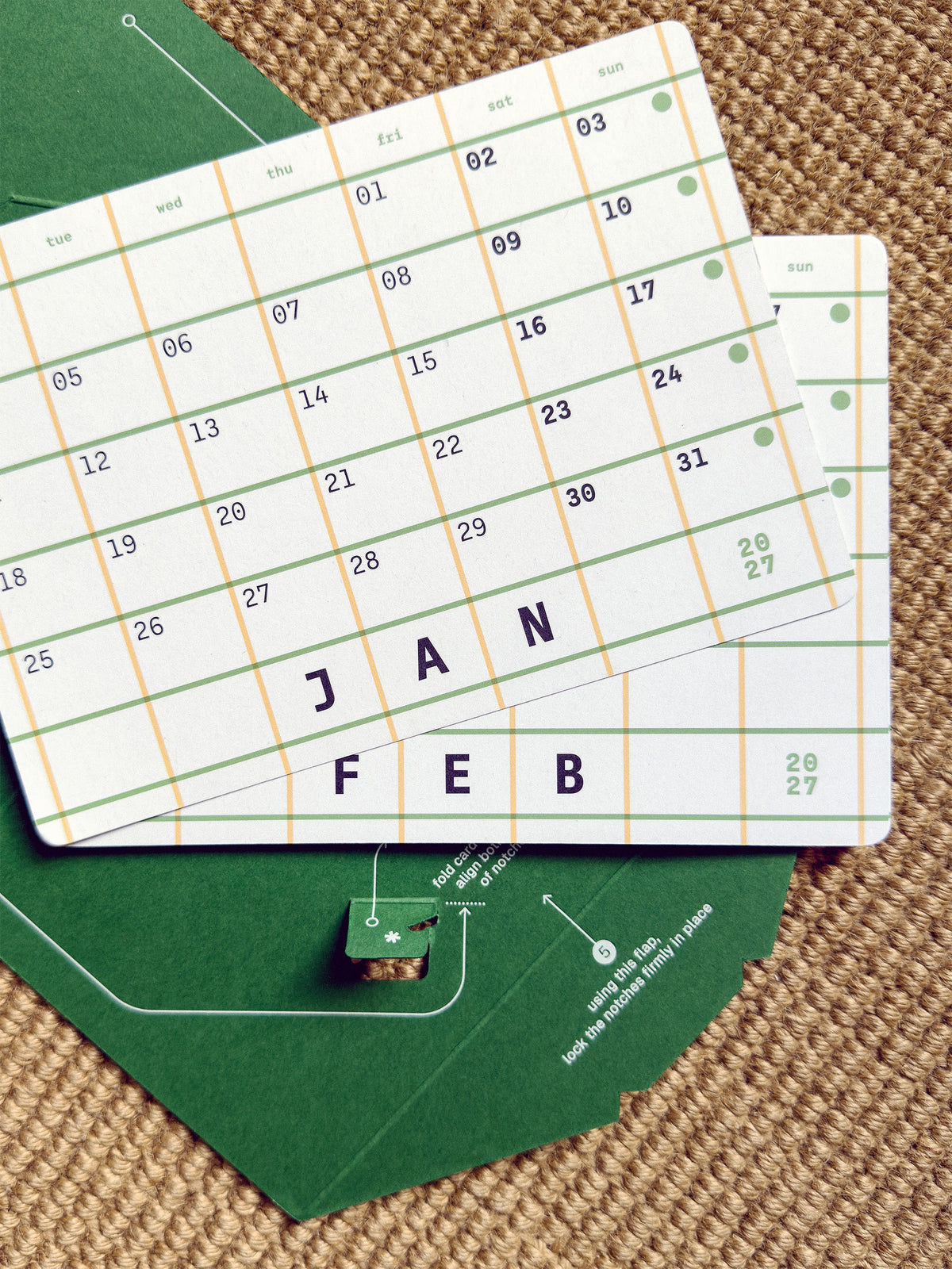 A close-up of a monthly calendar page with a grid layout and the months JAN, FEB, printed in large letters, placed on top of a green paper sheet over a woven surface.